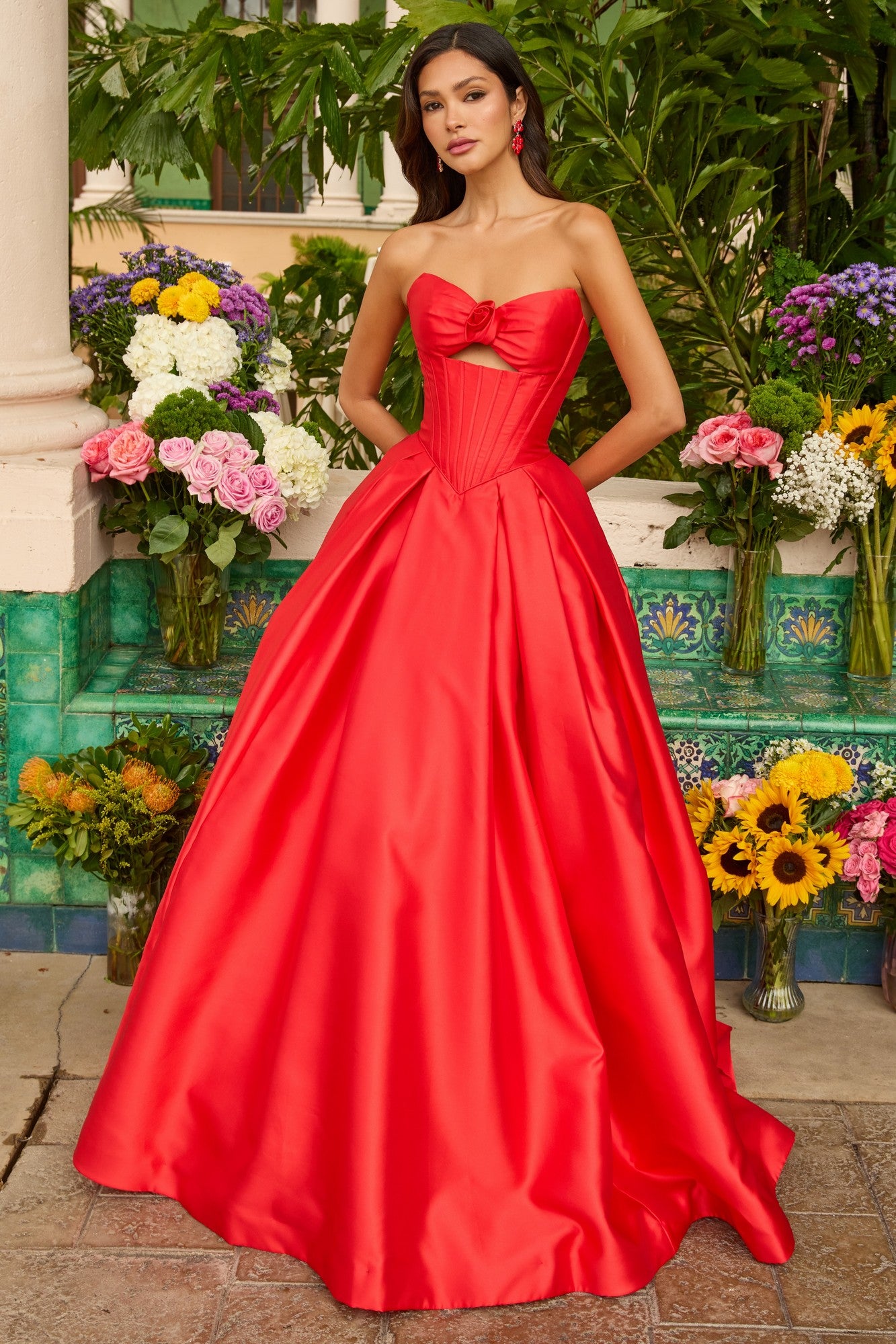 A woman in the Ava Presley 47223 Strapless Satin Prom Ball Gown—a bright red dress with a bow and cutout bodice—stands gracefully before vibrant outdoor flower arrangements.